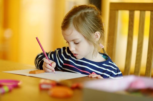 young girl sat at a dining room table with a book open and writing with a pencil.