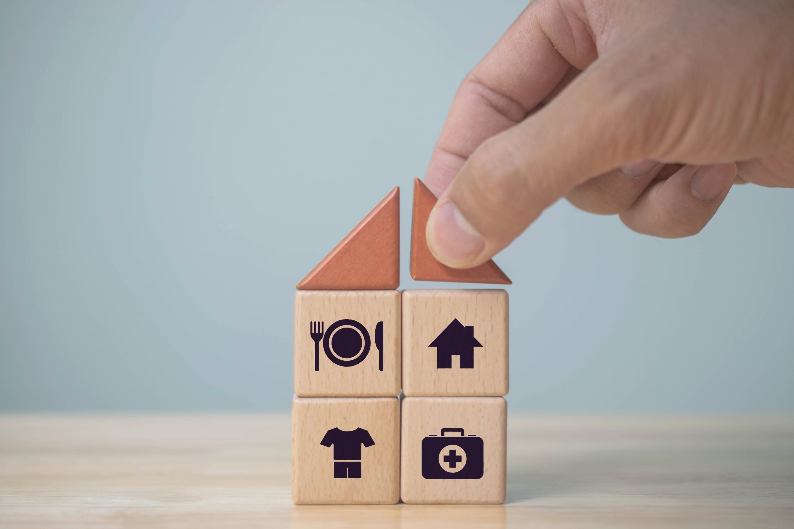 Hand placing a roof over four blocks making up a house, one with food, one with a home, one with clothing and the other with a camera representing access to technology.
