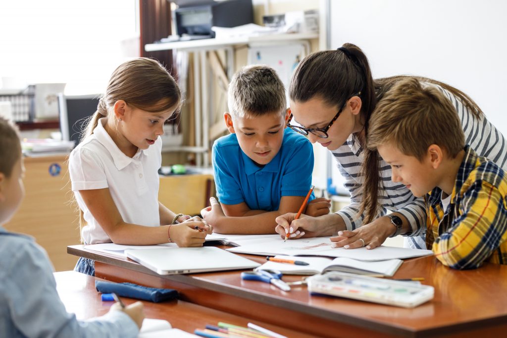 Female teacher helps school kids to finish they lesson. They are all sitting together at one desk.