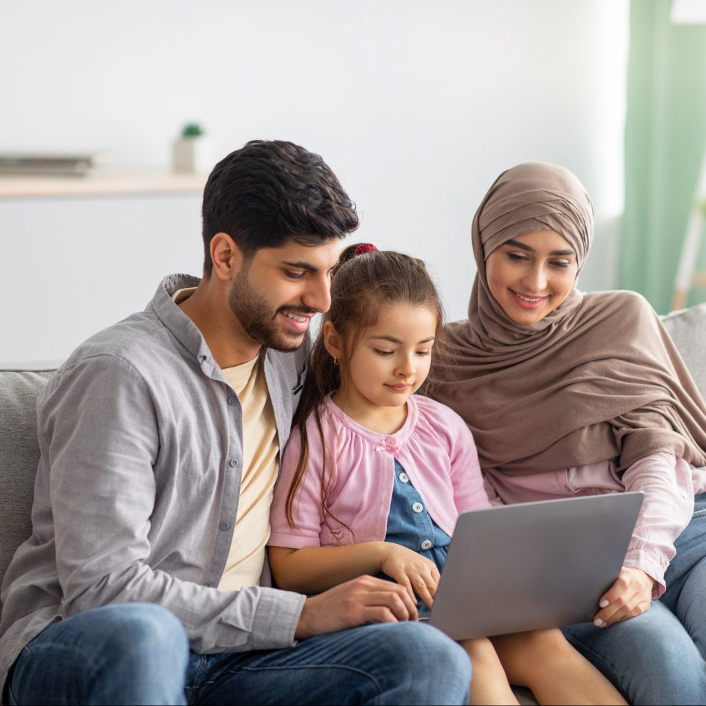 Young muslim parents and girl using laptop together at home, surfing internet or having video chat with someone, sittig on sofa. Happy arab man, child and woman in hijab staying home on isolation