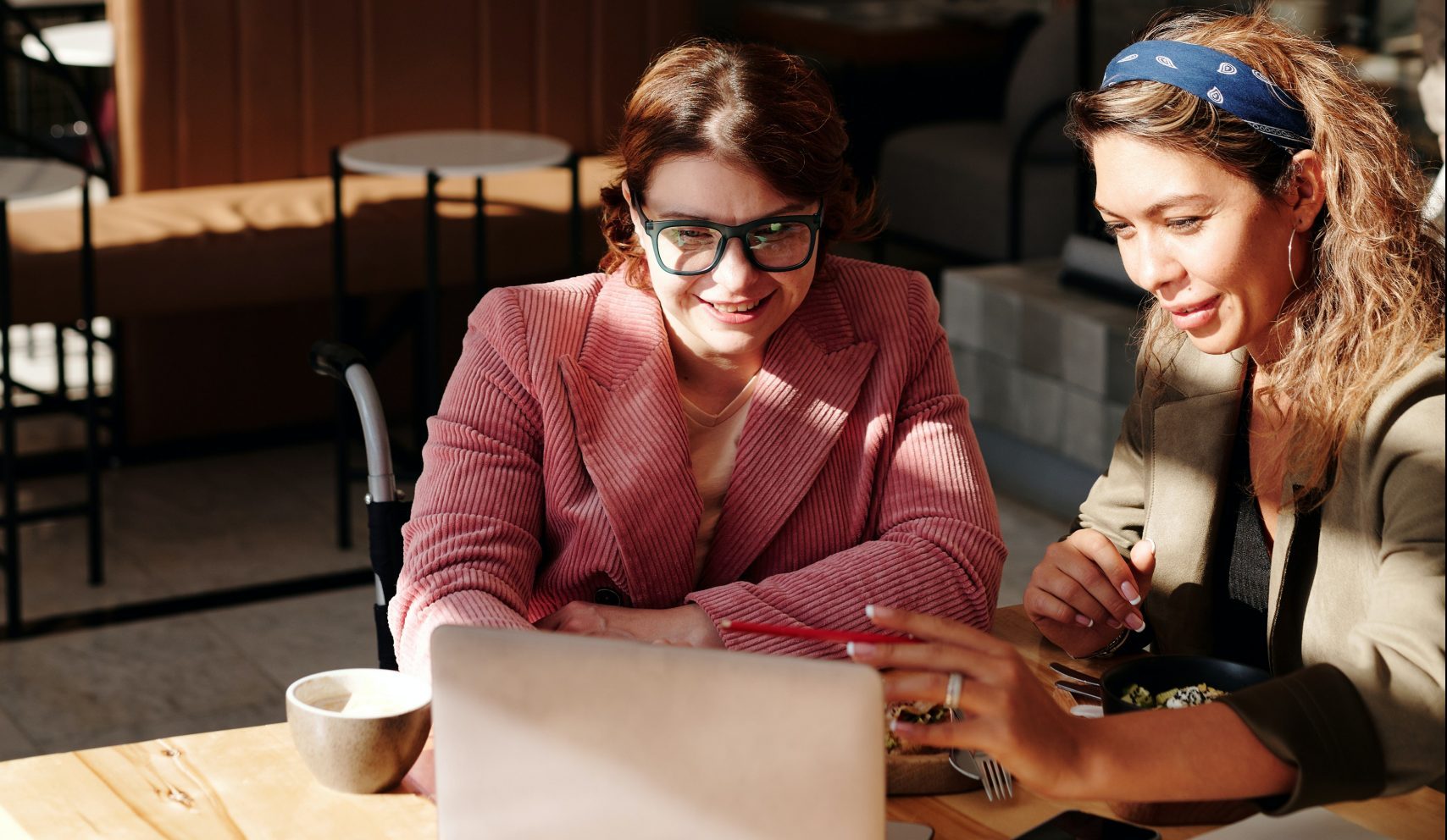Business woman dressed in a blazer in a wheelchair being trained in using a laptop at the table of a small cafe.
