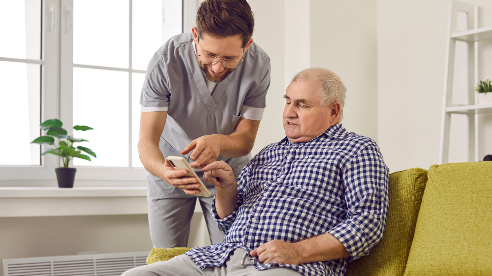 Young cheerful brunette nurse showing elderly man how to use smartphone. Nursing home service and patient support. Photo of caregiver teaching senior man to handle with mobile phone at home
