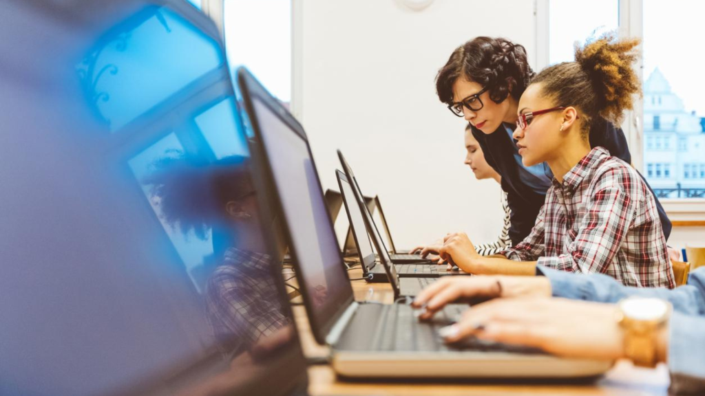 Tutor leaning over the shoulder of a young learner helping them whilst using a laptop
