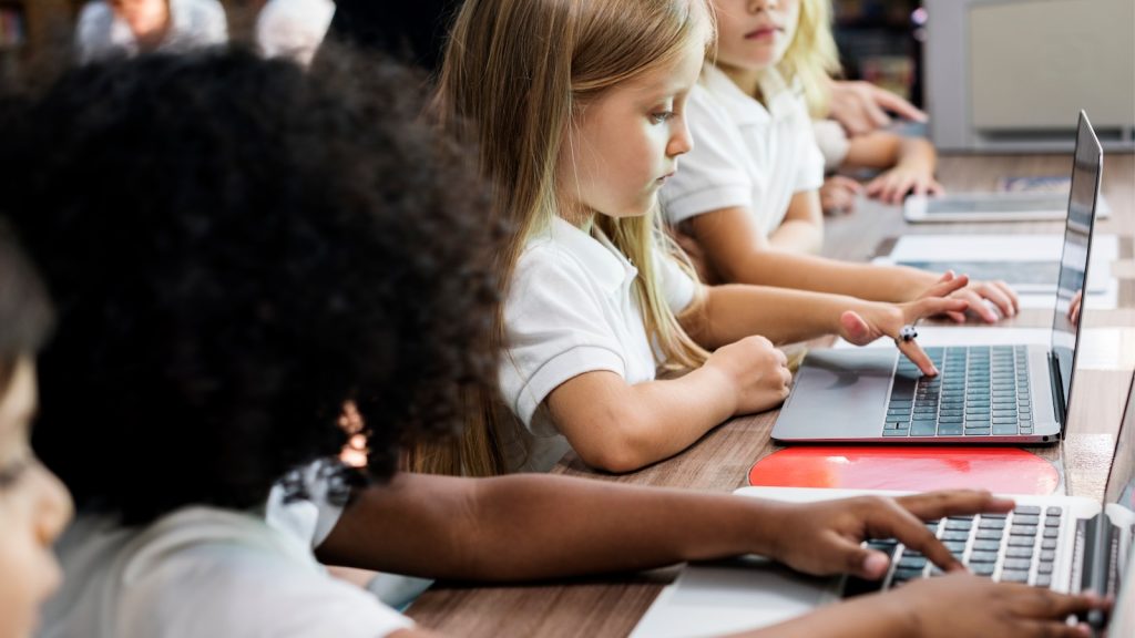 School-aged children at a desk working on laptops.